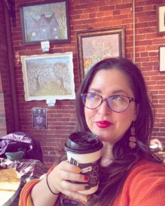 A woman enjoys a cup of coffee inside a Salem café, with local artwork featuring the House of the Seven Gables and autumn trees displayed on the brick wall behind her.