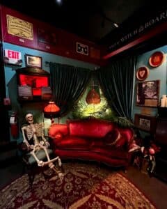 A quirky lounge setting featuring a red vintage sofa, a skeleton seated in an antique chair, and lush green drapes framing a glowing Salem Witch Board Museum emblem. The space is accented with eclectic decor, including framed artwork, a Victorian-style lamp, and a patterned rug.