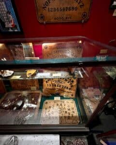 A glass display case at the Salem Witch Board Museum in Salem, Massachusetts, showcasing vintage Ouija boards, including "The Mystic Board" and "The Oracle," along with accompanying planchettes.