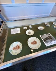 A glass display case inside the Salem Custom House in Salem, Massachusetts, showcasing souvenir ceramics featuring images of the Custom House and informational placards about their history.