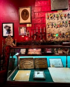 A section of the Salem Witch Board Museum in Salem, Massachusetts, showcasing vintage Ouija boards in a glass case, surrounded by antique photographs, candlesticks, and a decorative chair, set against a red backdrop.