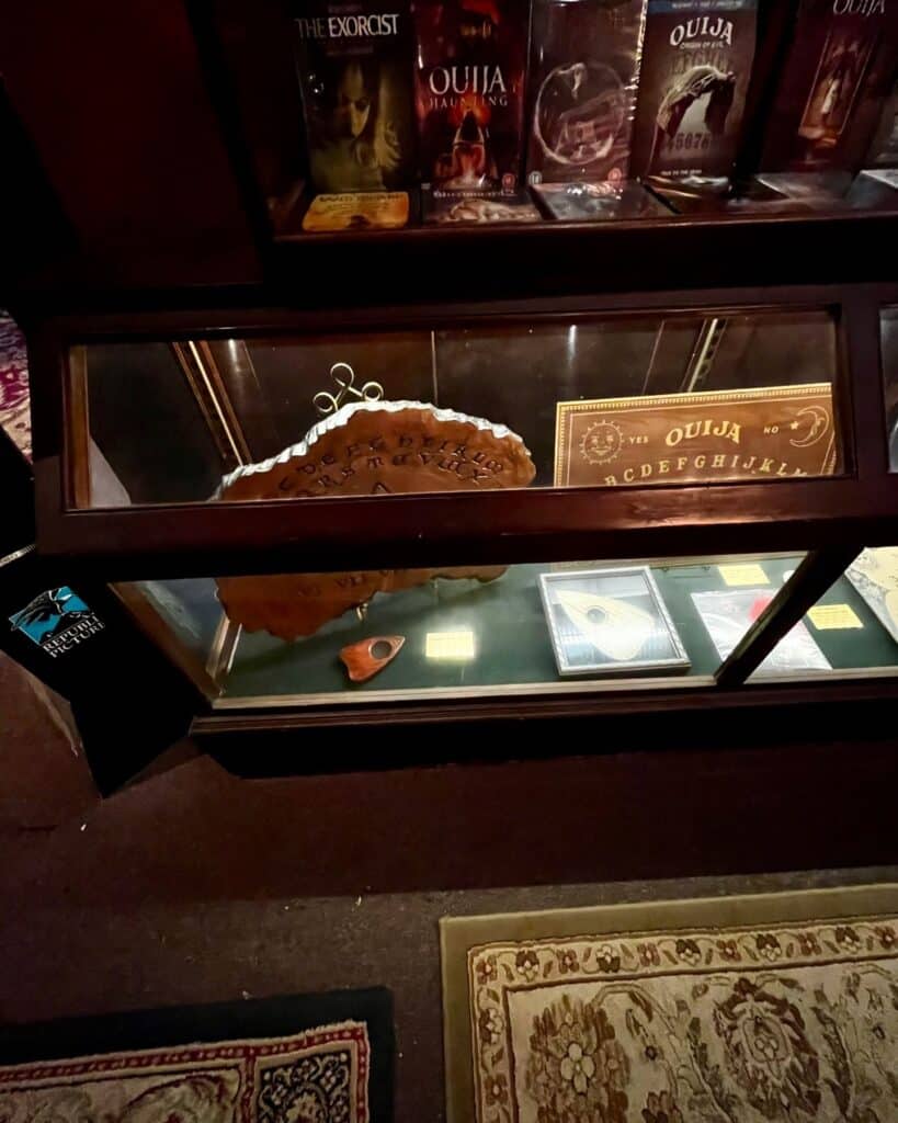 A glass display case at the Salem Witch Board Museum showcasing vintage Ouija boards and planchettes, accompanied by horror-themed DVDs like "The Exorcist" and "Ouija: Origin of Evil."
