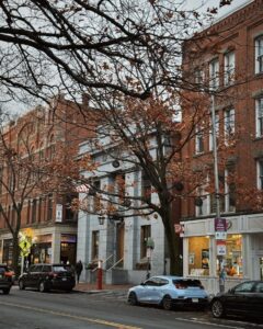 A street view in downtown Salem, Massachusetts, featuring historic brick buildings, shops, and a stone building with columns. A tree with autumn leaves partially frames the scene, with parked cars and pedestrians visible along the sidewalk.