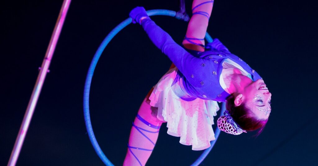 An aerial performer in Salem Massachusetts wearing a sparkling blue outfit, gracefully posing upside down on a hoop under colorful lights at the Frozen Fire Festival.
