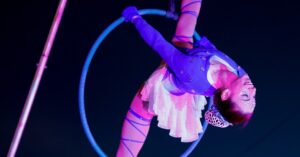 An aerial performer in Salem Massachusetts wearing a sparkling blue outfit, gracefully posing upside down on a hoop under colorful lights at the Frozen Fire Festival.