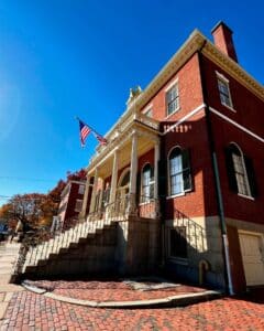 Angled view of the Salem Custom House in Salem, Massachusetts, showcasing its grand staircase, red brick exterior, and golden eagle under a clear blue sky.