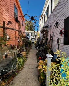 A festive alleyway in Salem, Massachusetts, decorated for Halloween with large hanging spider decorations, cobwebs, pumpkins, and gothic lanterns amidst vibrant orange and white buildings.