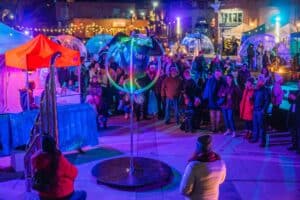 A glowing aerial hoop performance captivates the audience under colorful lights at the Frozen Fire Festival in Salem, Massachusetts, with illuminated igloos and festive booths in the background.
