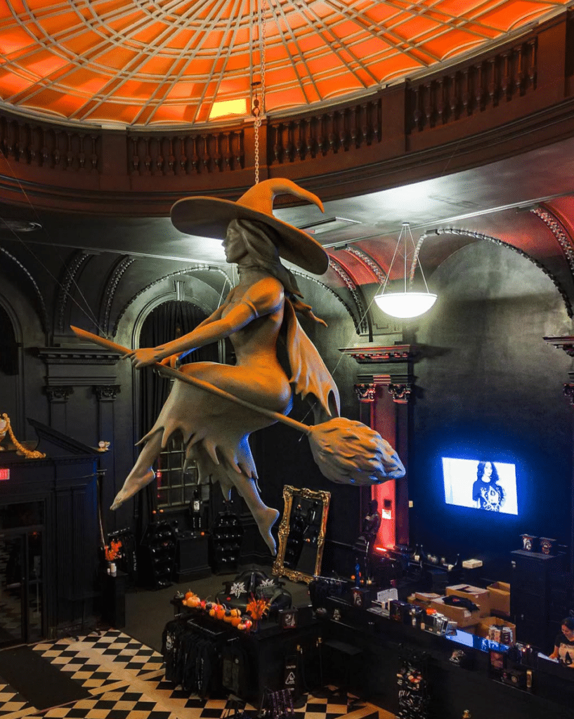  Inside Blackcraft Salem in Salem, Massachusetts, featuring a suspended witch sculpture riding a broom under an orange-lit dome ceiling, surrounded by gothic decor and merchandise displays.