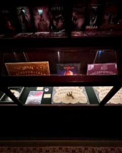 A dimly lit display case at the Salem Witch Board Museum in Salem, Massachusetts, showcasing Ouija boards, paranormal-themed book covers, and a mystical planchette.