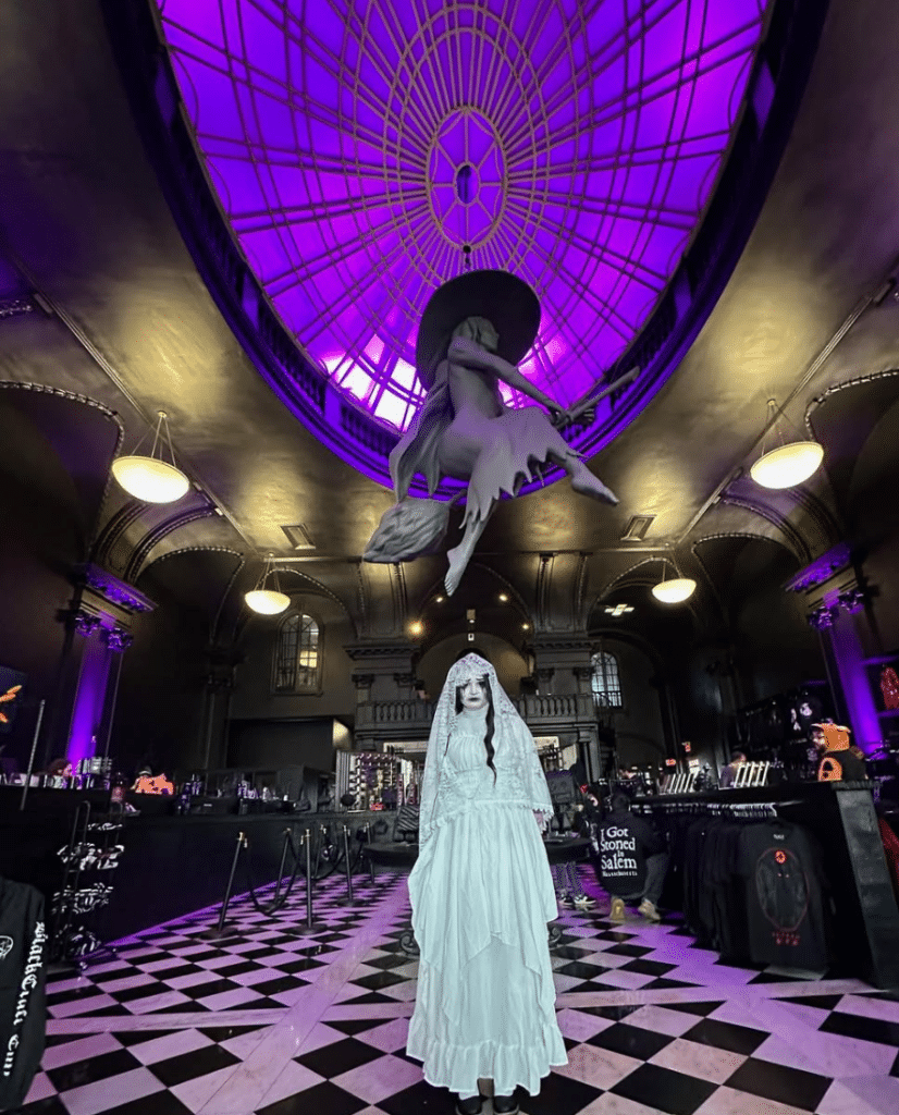 Gothic-themed interior of Blackcraft Cult store in Salem, Massachusetts, featuring a hauntingly dressed mannequin in a white veil and gown under a glowing purple dome with a suspended witch figure on a broomstick.