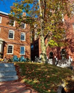 A serene autumn scene at The Merchant Hotel in Salem, Massachusetts, featuring red brick architecture, golden fall leaves, and cozy blue chairs on the lawn.