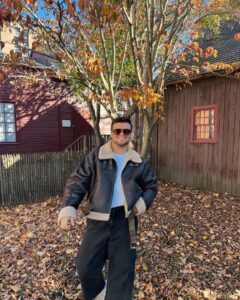 A person in a leather jacket stands in front of the Pickman House in Salem, Massachusetts, surrounded by autumn leaves and trees with fall foliage. The historic house in the background features red and brown wooden siding.