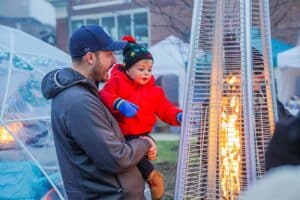 A father holds his young child as they warm up near a patio heater during the Salem Frozen Fire Festival in Salem, Massachusetts, with igloos and festival activities in the background.