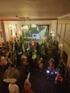 A Halloween ball at the Hawthorne Hotel with a crowd of costumed attendees, a DJ booth with colorful lights, and a grand ballroom setting with chandeliers.