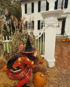 A woman in a witch costume crouches beside hay bales and pumpkins at the entrance of Ropes Mansion in Salem, Massachusetts. She wears a black witch hat adorned with colorful pom-poms and an orange jacket with sun and moon designs.
