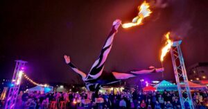 A skilled performer in Salem Massachusetts balancing upside down on hand supports at the Frozen Fire Festival, holding flaming torches with outstretched limbs beneath colorful evening lights.