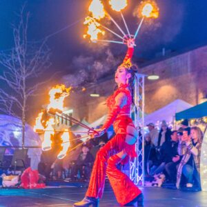 A performer in a vibrant red outfit twirls flaming props during a dramatic nighttime show, captivating an audience in Salem, Massachusetts.