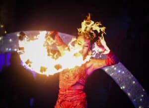 A fire dancer in an ornate red costume performs with fiery props, surrounded by flames and glowing lights during a dramatic nighttime show in Salem, Massachusetts.