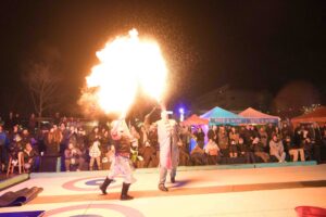 Two performers breathe fire, captivating a bundled-up audience under the night sky during a lively outdoor winter event in Salem, Massachusetts.