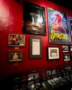 A red wall in the Salem Witch Board Museum features large posters of the films "Ouija" and "Spiritika," alongside framed photographs and memorabilia related to Ouija boards. Below, a glass case displays DVDs and books on the subject.