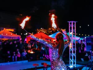 A performer in a reflective costume skillfully twirls flaming batons in front of a captivated audience at a nighttime event in Salem, Massachusetts.