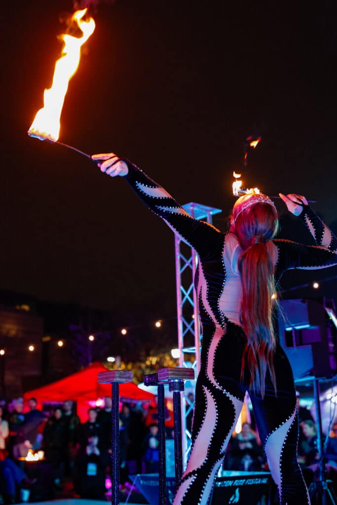 A performer in a sleek black costume with shimmering details wields flaming batons during a captivating nighttime show in Salem, Massachusetts.