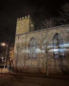 The First Church of Salem, a historic stone building with Gothic-style arched windows and a square tower, illuminated at night. A leafless tree stands in front, with shadows cast on the building.