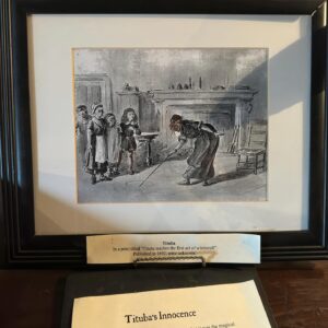 A framed illustration depicting Tituba, a woman accused during the Salem witch trials, teaching a group of children in a colonial-era room. The image shows Tituba bending down, drawing on the floor with a stick, while several children stand and watch. Below the framed picture is a plaque titled "Tituba's Innocence" along with an additional description of the artwork.