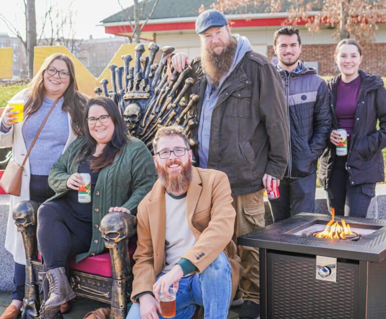 A group of friends poses around a medieval-themed throne at the Salem Frozen Fire Festival in Salem, Massachusetts, enjoying drinks and a nearby fire pit.