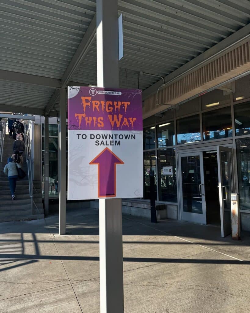 A sign reading "Fright This Way to Downtown Salem" with a purple arrow stands at the Salem train station. The sign has a spooky design, and people are seen walking up the stairs nearby.
