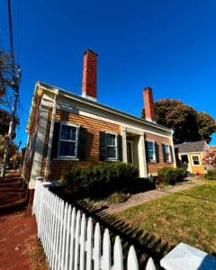 A front angled view of the Jonathan Whipple House in Salem, Massachusetts, showing its yellow siding, white trim, red brick chimneys, and charming white picket fence under a bright blue sky.