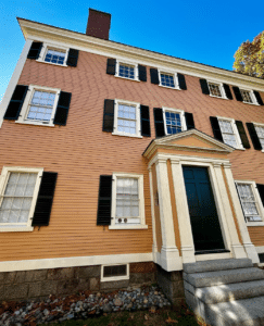The front view of the Hawkes House in Salem, Massachusetts, showcasing its peach clapboard siding, black shutters, white trim, and an elegant entryway with a dark green door under a clear blue sky.