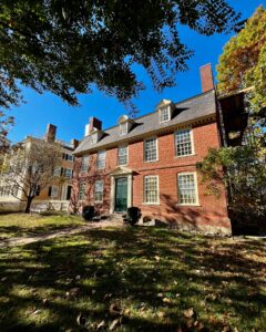 The Derby House in Salem, Massachusetts, showcasing its brick Georgian architecture, symmetrical windows, a central green door, and a tree-lined yard under a bright blue sky.