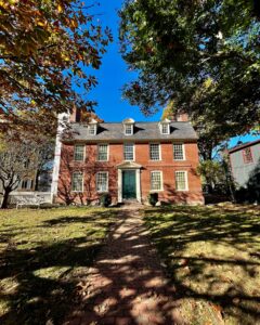 The Derby House in Salem, Massachusetts, featuring its red brick Georgian architecture, a green door, symmetrical windows, and a brick path leading through a tree-lined yard under a bright blue sky.