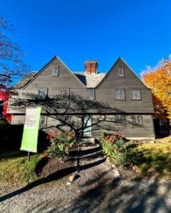 The front facade of the John Ward House in Salem, Massachusetts, featuring a dark wooden exterior, lattice windows, and a central chimney, surrounded by blooming red flowers and shaded by tree branches under a vibrant blue sky.
