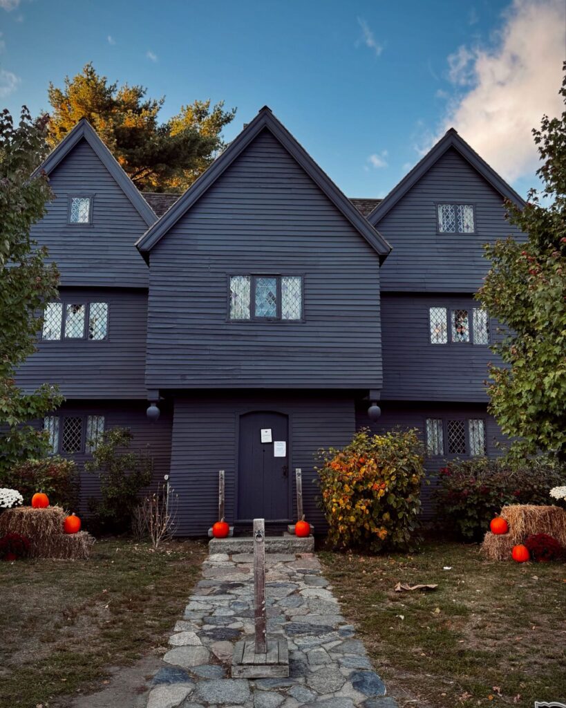 The Salem Witch House, painted dark gray, with its iconic peaked rooflines against a clear sky. The entrance is adorned with orange pumpkins and hay bales, creating a festive autumn scene, with a stone pathway leading up to the door.