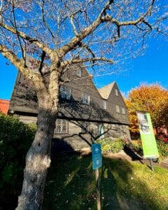 The front facade of the John Ward House in Salem, Massachusetts, featuring its dark wood exterior and diamond-paned windows, framed by a leafless tree with sprawling branches under a vibrant blue sky.