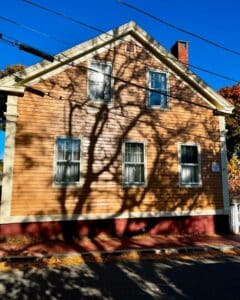 The front facade of the Jonathan Whipple House in Salem, Massachusetts, featuring its yellow siding, four large windows, and dramatic tree shadows under a bright blue sky.