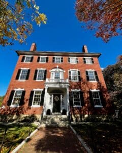 The William Pickman House in Salem, Massachusetts, a historic red brick building with black shutters and a grand white entryway, framed by autumn trees and a clear blue sky.