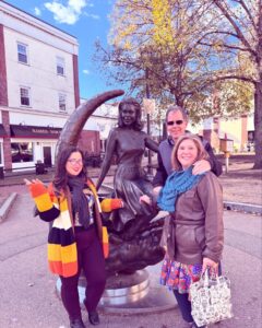 Three people pose with the famous Bewitched statue in Salem, Massachusetts, which features Samantha Stephens from the classic TV show. The statue is surrounded by fall trees and a downtown backdrop.