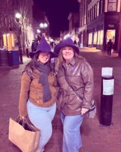 Two people wearing purple witch hats pose on Essex Street in Salem, Massachusetts, at night with historic buildings and streetlights in the background.