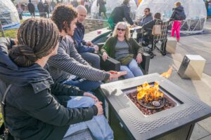 A group of people sits around a fire pit, enjoying the Salem Frozen Fire Festival's cozy outdoor setting with igloos and other festivalgoers in the background in Salem, Massachusetts.