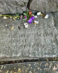 Memorial stone for George Jacobs at the Salem Witch Trials Memorial in Salem Massachusetts, engraved with his name, the date August 19, 1692, and surrounded by vibrant flowers and seashell offerings left by visitors.