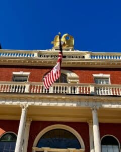 The Salem Custom House in Salem, Massachusetts, showcasing a golden eagle perched atop the building, white columns, detailed architectural features, and an American flag waving under a bright blue sky.