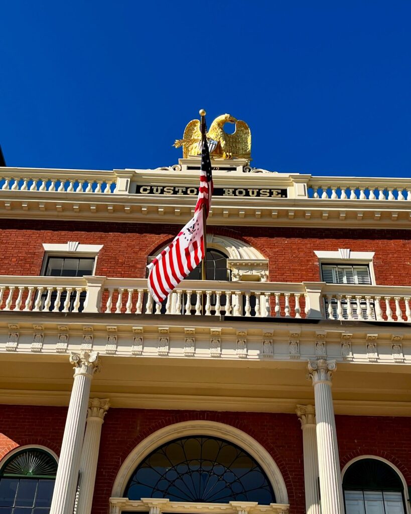 The Salem Custom House in Salem, Massachusetts, showcasing a golden eagle perched atop the building, white columns, detailed architectural features, and an American flag waving under a bright blue sky.