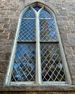 A close-up of a tall, arched window at the First Church of Salem in Massachusetts, featuring intricate diamond-patterned glass panes set in a stone frame, reflecting trees and the sky.