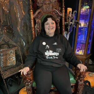 A person dressed in gothic attire sits in an ornate wooden throne-like chair. The background is filled with eerie Halloween decor, including cobwebbed candleholders, pumpkins, and an antique chest. The ambiance has a dark and mysterious vibe, with intricate designs and dramatic lighting adding to the gothic atmosphere.
