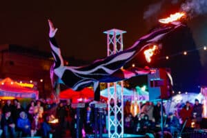 An acrobat in a sleek black costume performs a one-handed balance on elevated stands while holding flaming props, surrounded by a vibrant crowd in Salem, Massachusetts.