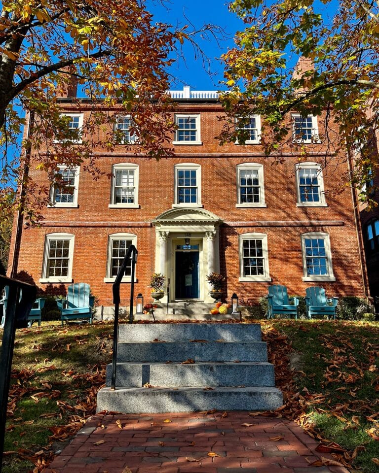The Merchant Hotel in Salem, Massachusetts, features a stately brick facade, adorned with a white columned entrance, autumn leaves on the lawn, and blue Adirondack chairs under the colorful trees.